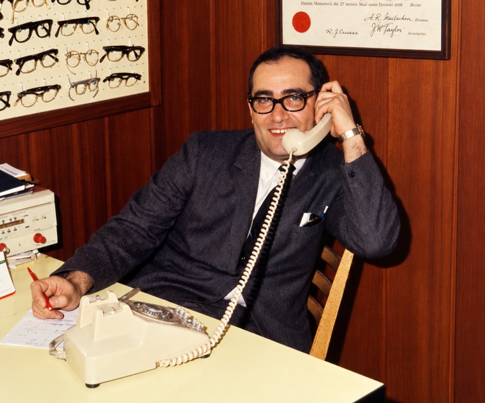 Photo d'archive en couleur : un homme en complet et lunettes à monture noire, souriant, le combiné d'un téléphone à cadran à l'oreille, assis à un bureau devant un présentoir de montures et un diplôme encadré.