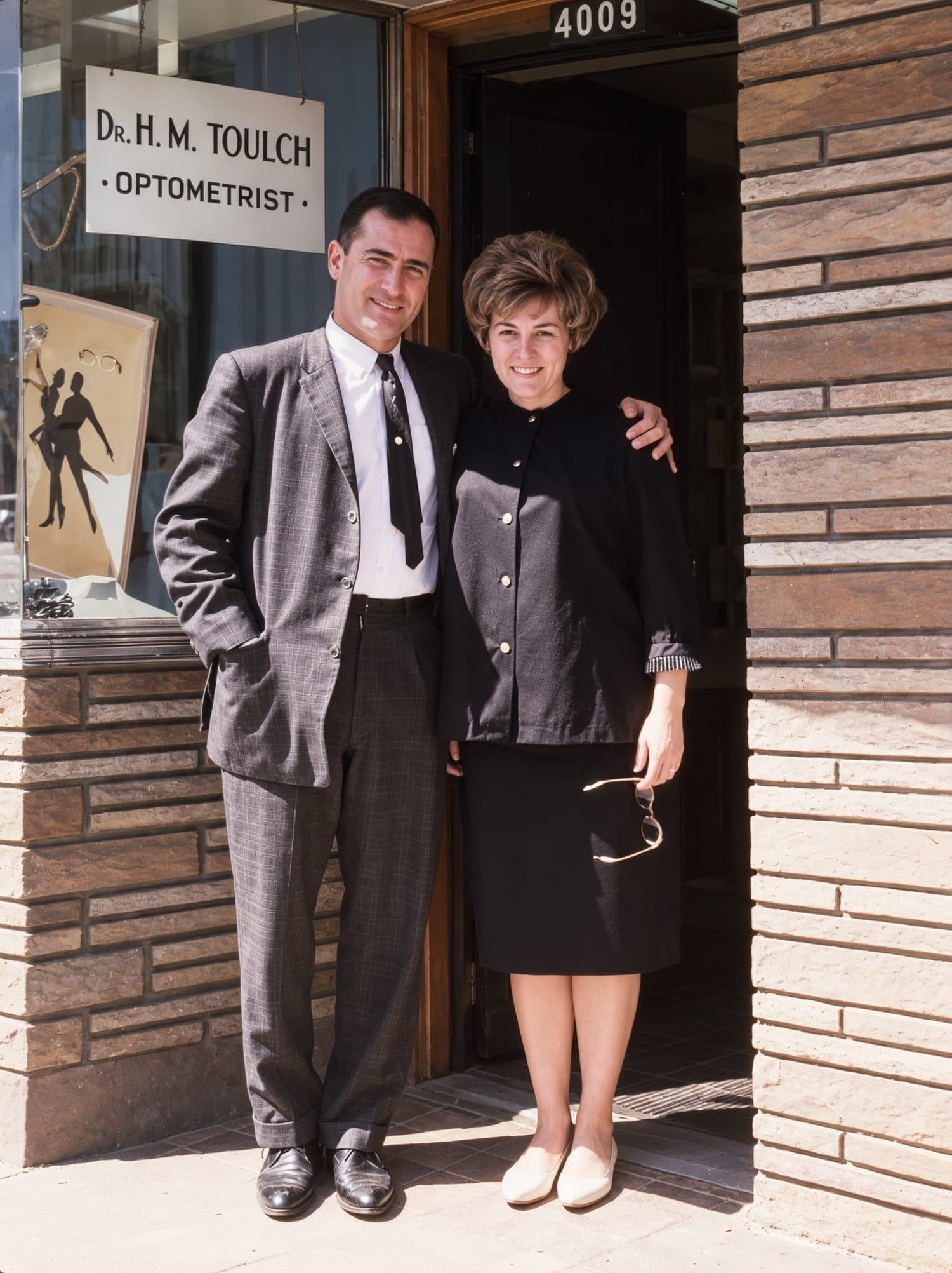 Photo d'archive en couleur : Harry et Libby Toulch sur le seuil de leur premier local du boulevard Saint-Laurent, sous l'enseigne « Dr. H.M. Toulch, Optometrist ».