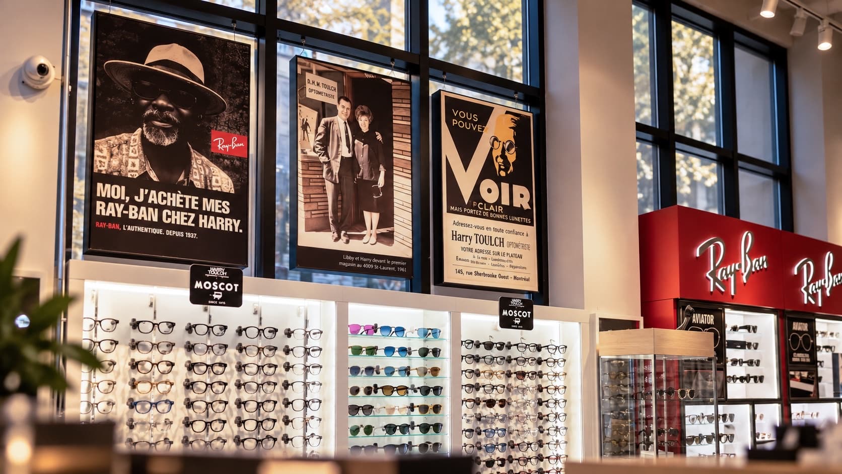 Le mur d'affiches de la boutique Harry Toulch : une affiche Ray-Ban, la photo de Harry et Libby devant leur premier magasin en 1961, et une affiche d'époque « Voir clair », au-dessus des présentoirs Moscot et du comptoir Ray-Ban.