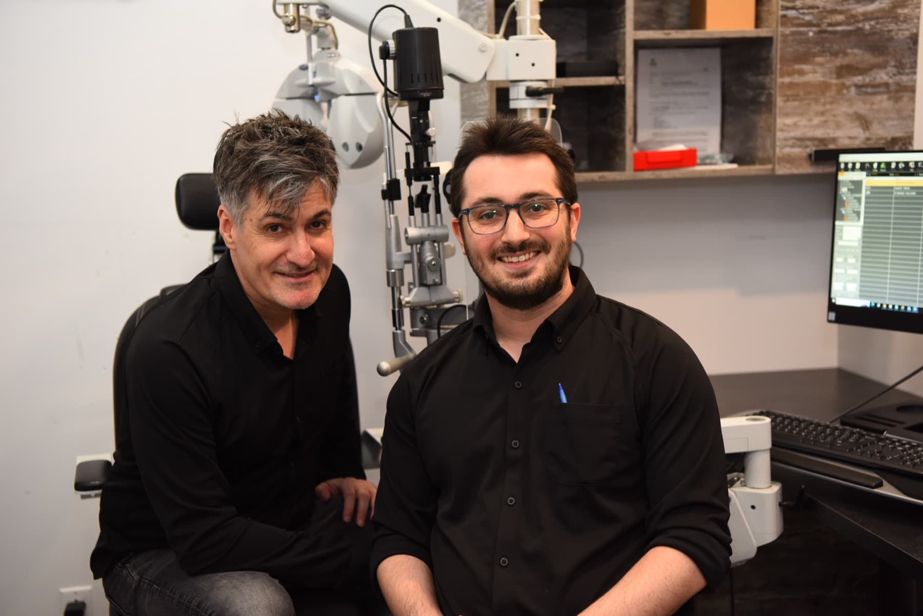 Les optométristes Michael Toulch et son fils Jonah Toulch, souriants, dans une salle d'examen, devant une lampe à fente.