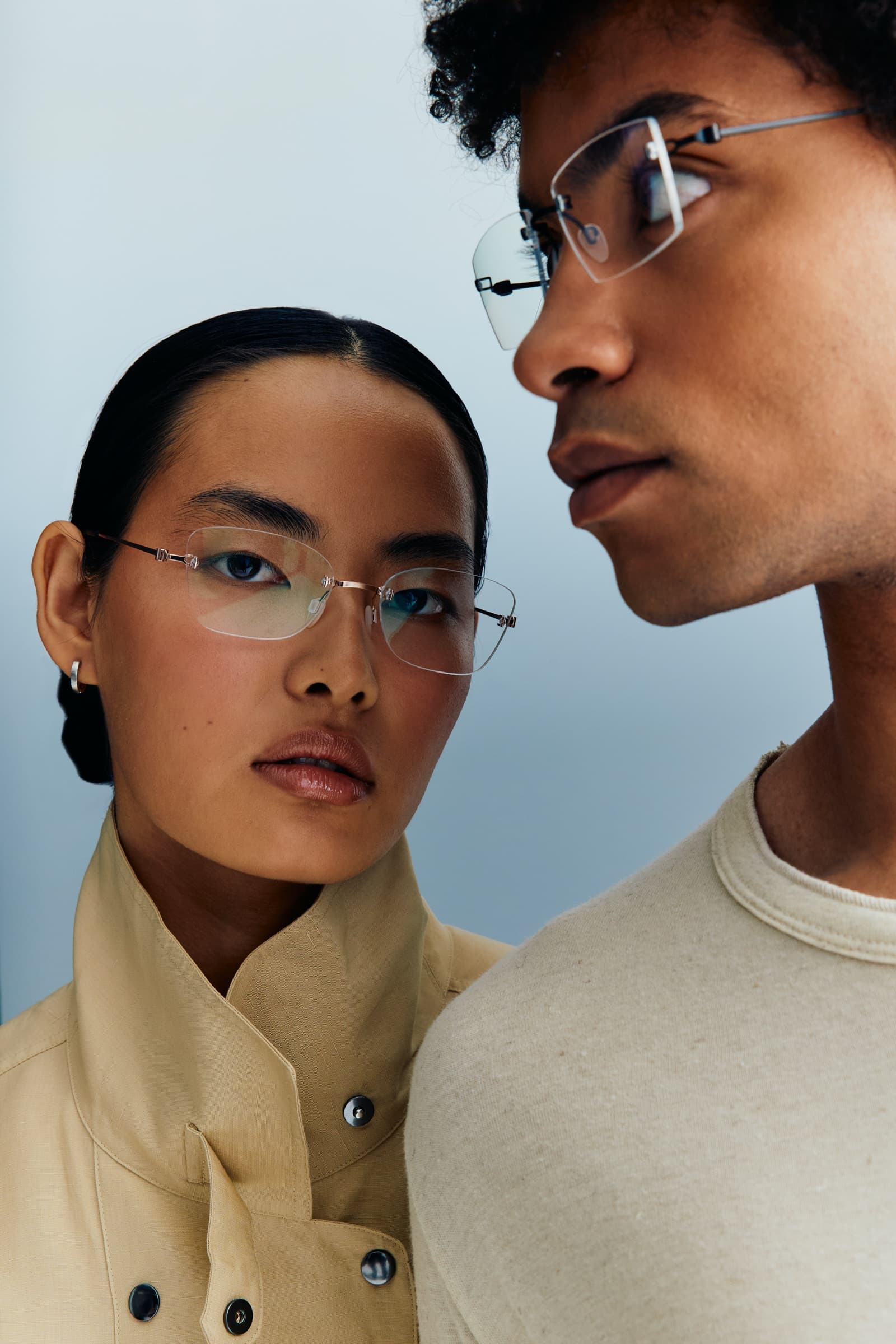 A woman and a man wearing Toulch Rimless frames: she looks at the camera in a rose-gold pair, he is in profile in a darker pair, against a pale blue-grey background.